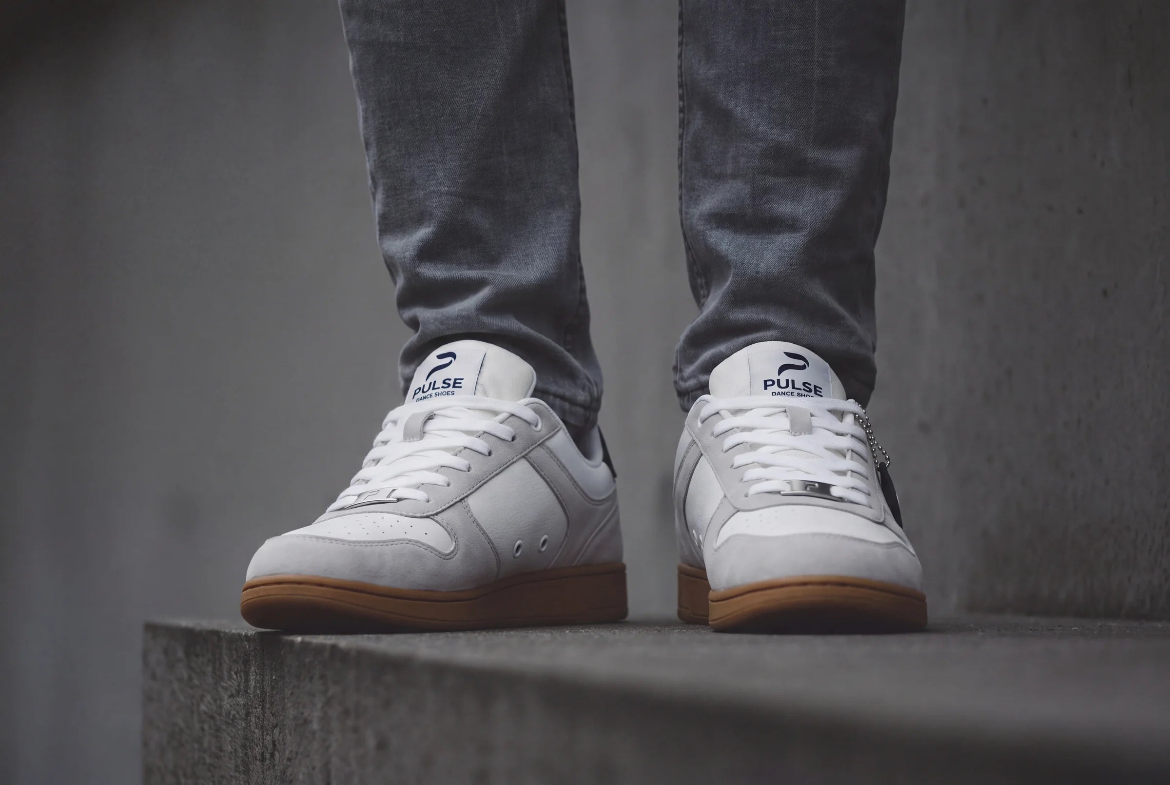 Feet in white dance sneakers with tan PU soles and grey jeans, standing on a concrete ledge
